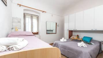 A bedroom with two beds, a wooden headboard, a window with curtains, a mirror, a cross on the wall, and a small table with towels and a tray.