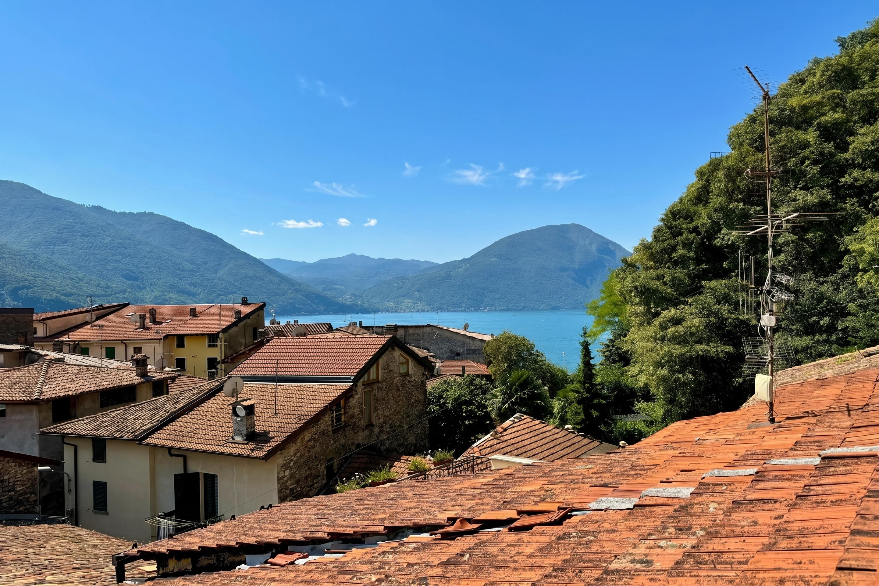 A coastal town with terracotta-roofed buildings, a clear blue sky, and a view of the sea and mountains in the background.