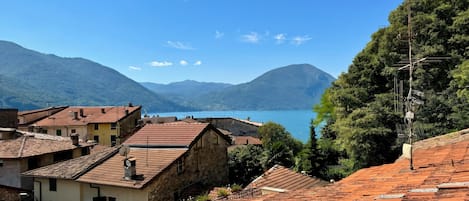 A coastal town with terracotta-roofed buildings, a clear blue sky, and a view of the sea and mountains in the background.