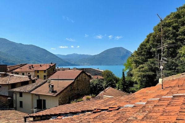 A coastal town with terracotta-roofed buildings, a clear blue sky, and a view of the sea and mountains in the background.