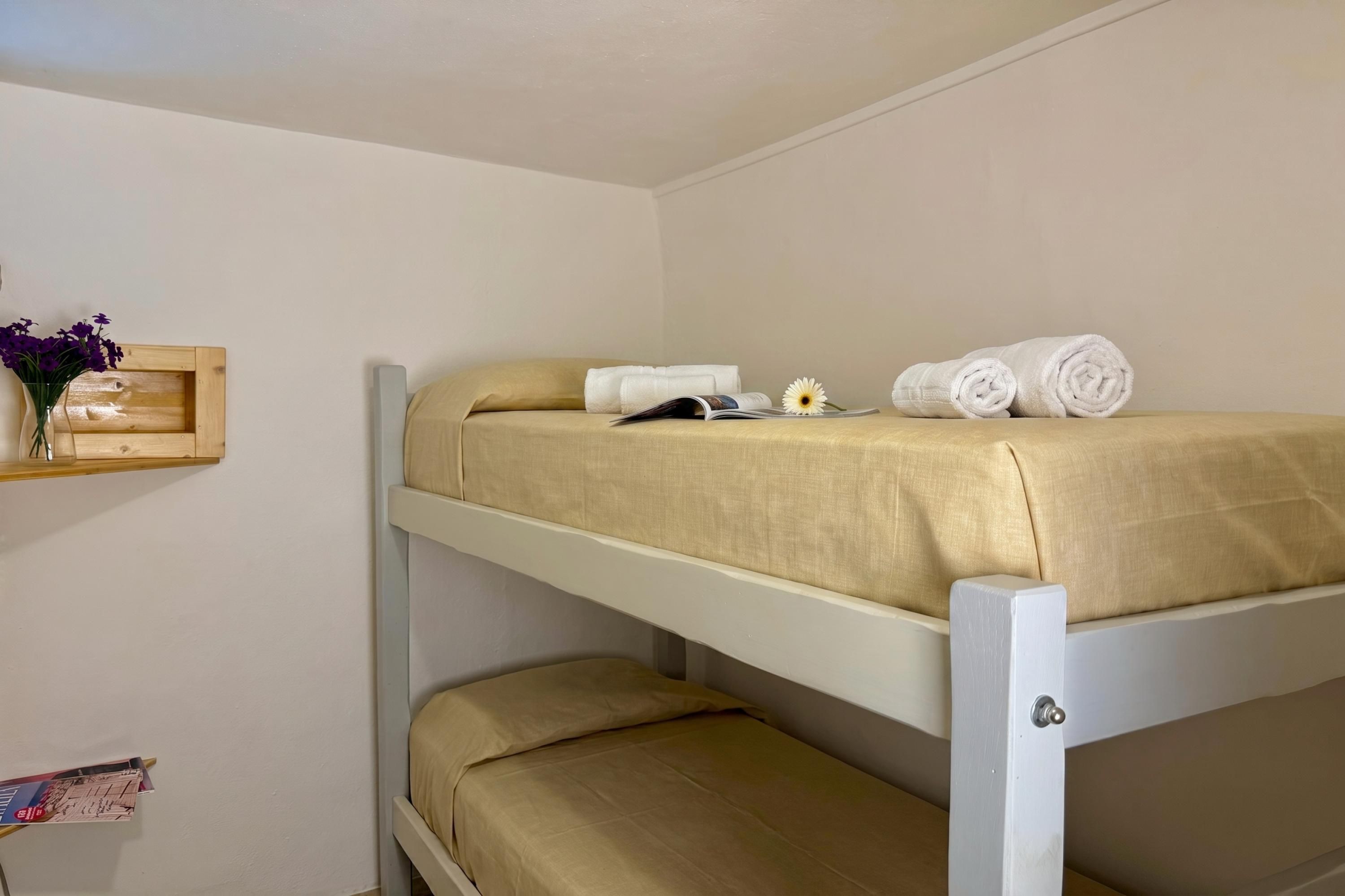 A bunk bed with a beige mattress, a wooden shelf with a vase of flowers, and a book on the lower bunk.