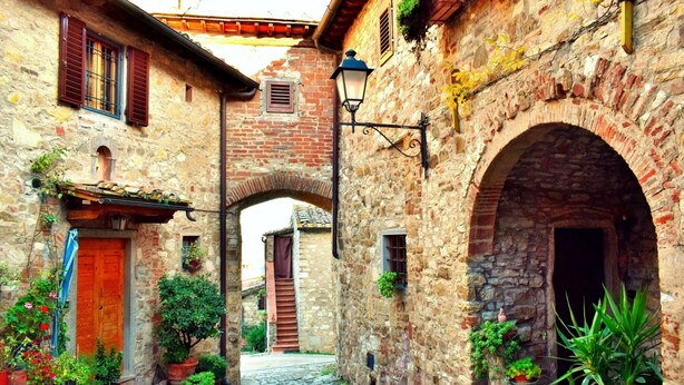 A narrow cobblestone street flanked by stone buildings with arched doorways and wooden shutters.