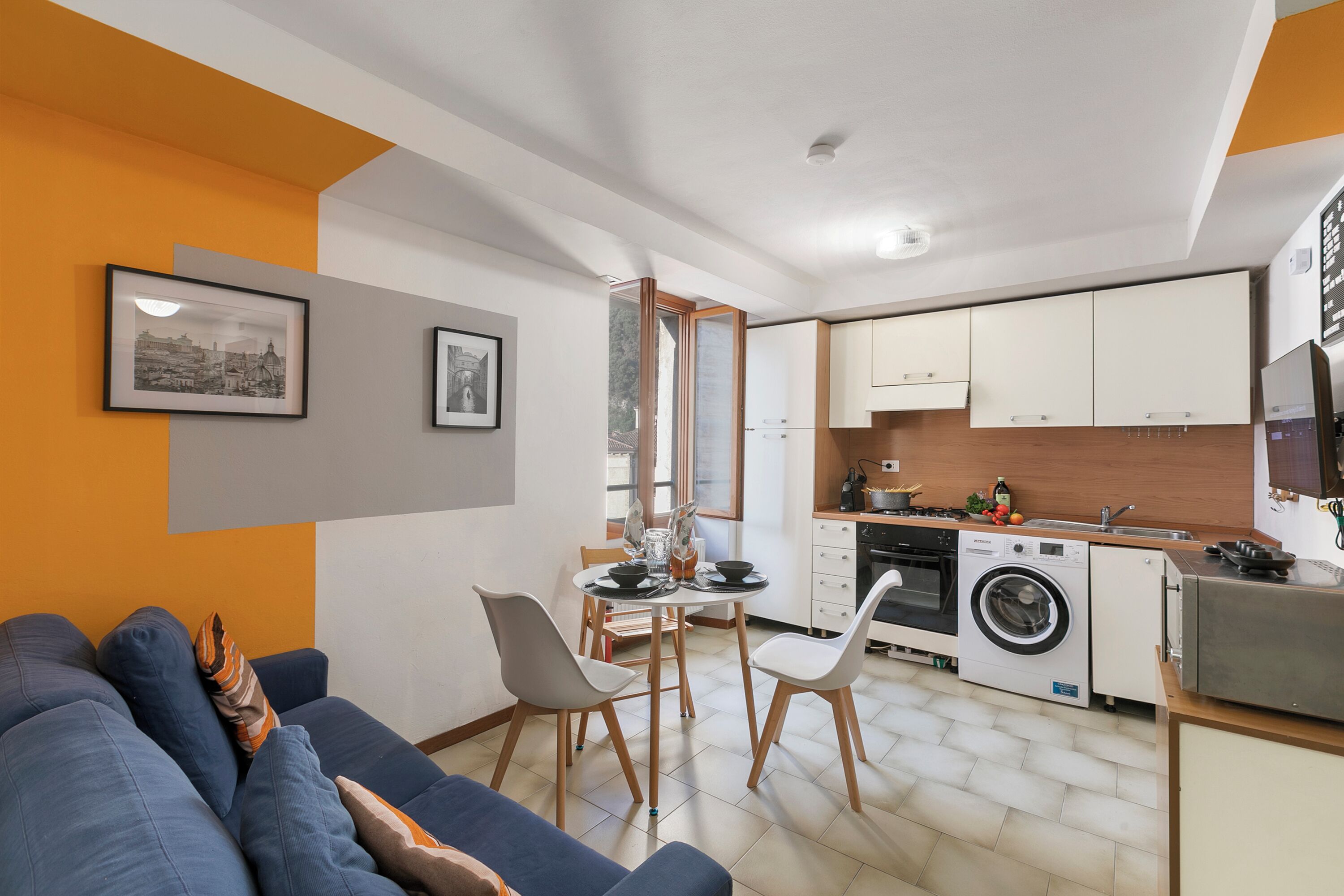 A compact living space with a sofa, dining table, chairs, a washing machine, and a kitchenette.