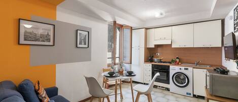 A compact living space with a sofa, dining table, chairs, a washing machine, and a kitchenette.