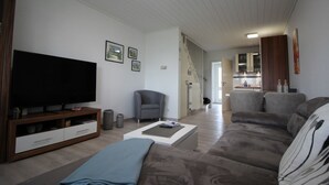 A living room with a flat-screen TV, a grey armchair, a sofa, and a coffee table. There is a staircase leading to an upper floor. - Ferienhaus in Busum fur 4 Personen (Büsum)
