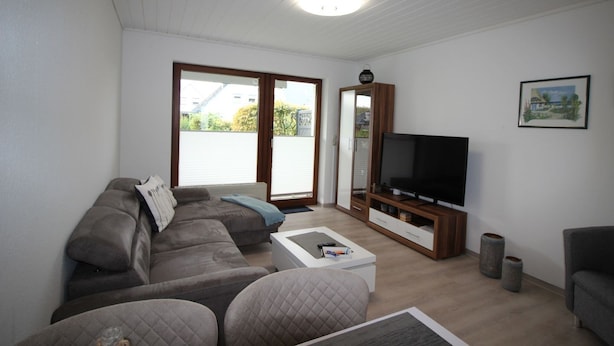 A modern living room with a sofa, coffee table, TV, and a sliding glass door leading to a balcony. - Ferienhaus in Busum fur 4 Personen (Büsum)