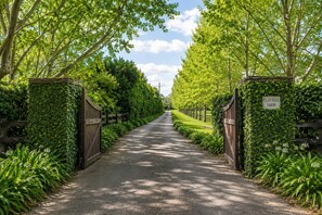 A paved pathway flanked by lush greenery and a wooden gate, leading to a property named 'Flat Hill Farm.'