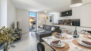 A modern living room with a sofa, dining table set for six, and a kitchen area with appliances. - Corte di Ascona Apartments (Ascona)