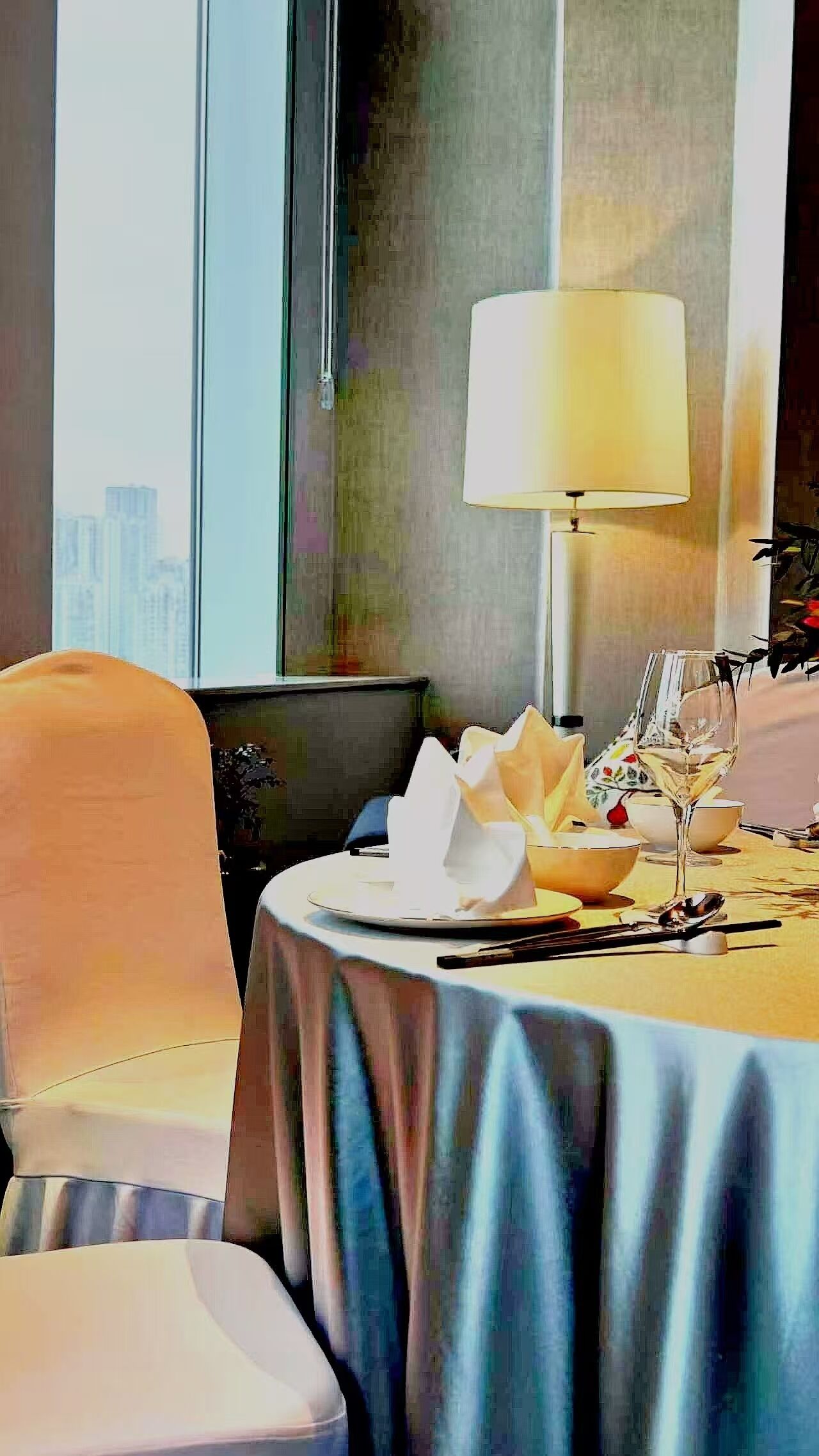 A hotel room with a dining table set for two, a lamp, and a view of the cityscape through large windows.