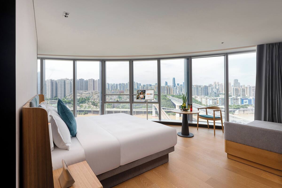 Una habitación de hotel moderna con una cama grande, una mesita con una silla y vistas a un paisaje urbano a través de amplios ventanales.