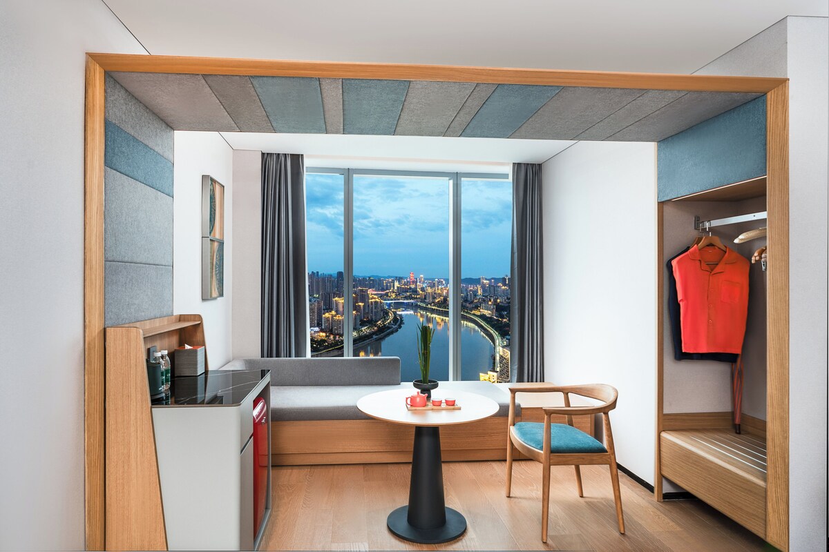 A modern hotel room with a large window, a round table, a sofa, a chair, a small kitchen area, and a wardrobe.