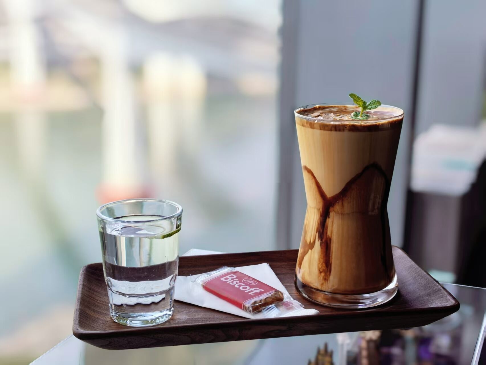 A tray with a tall glass of iced coffee, a shot glass, and a biscuit packet on a table by a window.