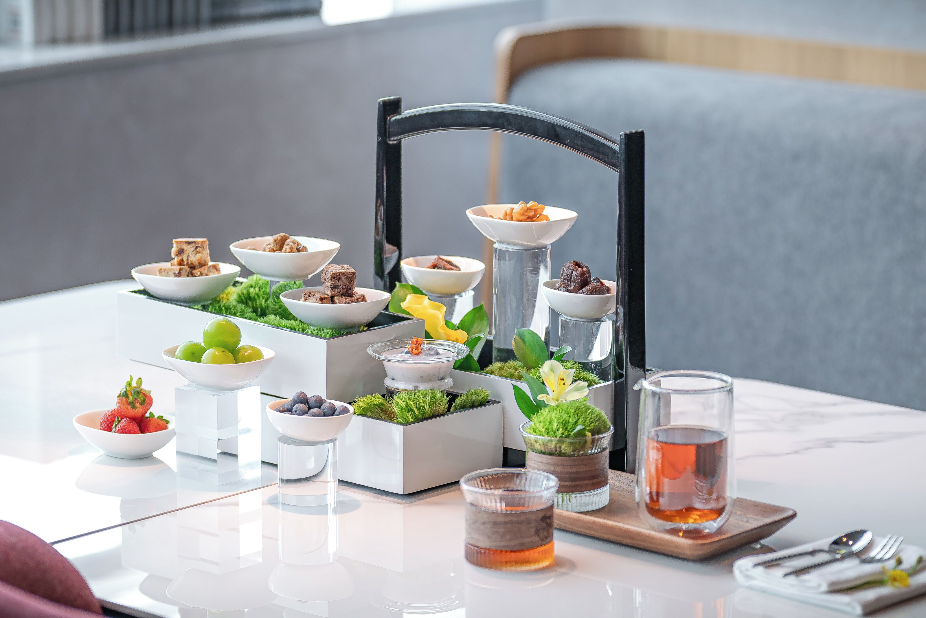 Una mesa de comedor con diversos alimentos y bebidas, incluyendo tazones de frutas, una taza de té y una bandeja con cubiertos.
