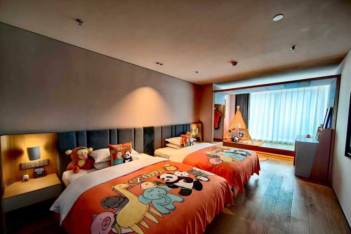 Habitación de hotel con dos camas, cada una con una colcha de motivos animales y colores vivos. Se ve una ventana con cortinas, una mesita de noche con lámpara y un escritorio pequeño con una computadora.
