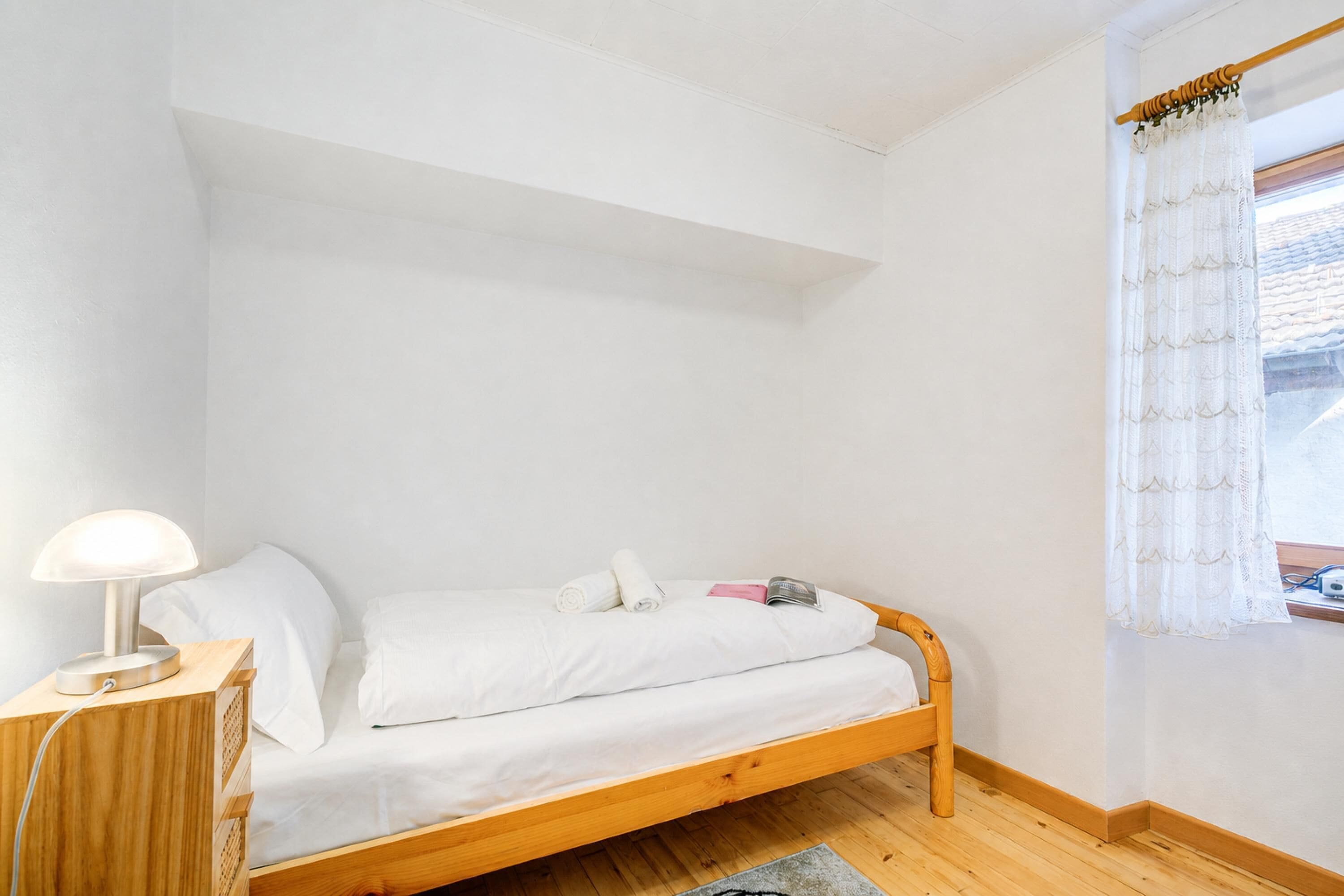 A single bed with a wooden frame, white bedding, a bedside lamp, and a window with sheer curtains.