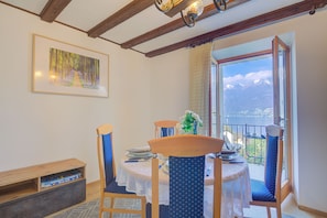 Un comedor con una mesa preparada para dos personas, sillas con tapicería azul, un banco de madera, un cuadro enmarcado en la pared y vistas a montañas a través de un ventanal abierto.