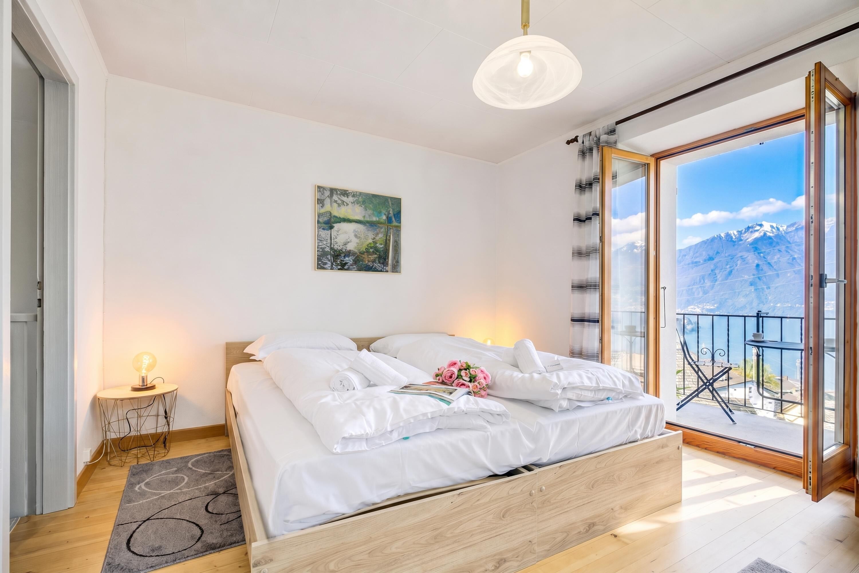 Ein Schlafzimmer mit einem Bett, einem Nachttisch, einer Lampe, einem Gemälde und Blick auf Berge vom Balkon aus.