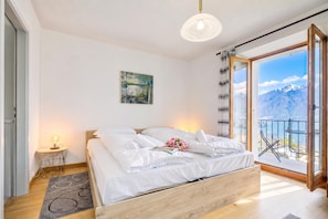 Un dormitorio con cama, mesita de noche, lámpara, un cuadro y vistas a las montañas desde un balcón.