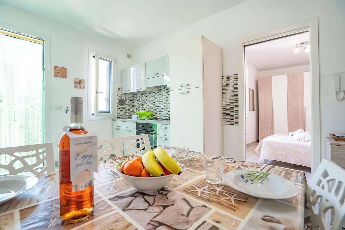 Una cocina moderna con un lavamanos verde, un comedor con una mesa preparada para dos personas y un dormitorio con una cama y una mesita de noche.