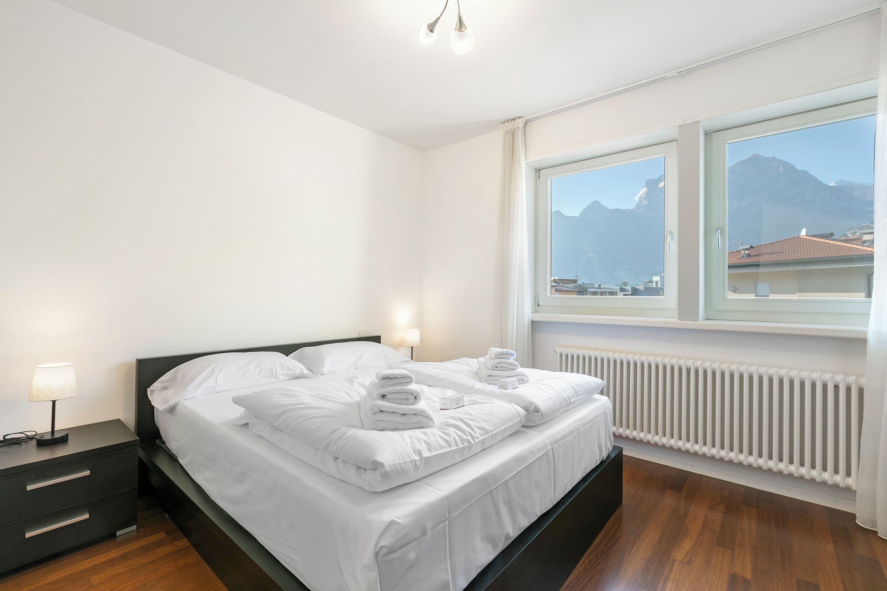 A neatly made bed with white linens and towels, a bedside table with a lamp, and a view of mountains through a window.