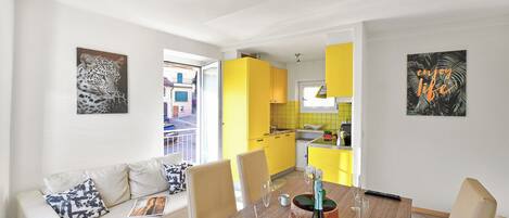 A modern living room with a sofa, dining table, and chairs. There is a kitchen in the background with yellow cabinets.