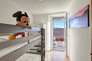 Un lit superposé avec un jouet en peluche sur le lit du haut, une porte-fenêtre coulissante donnant sur un espace extérieur avec une chaise, et un tableau accroché au mur.