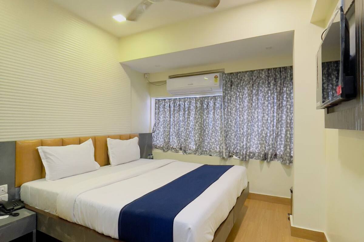 A hotel room with a bed, a ceiling fan, an air conditioning unit, a TV mounted on the wall, and a window with curtains.