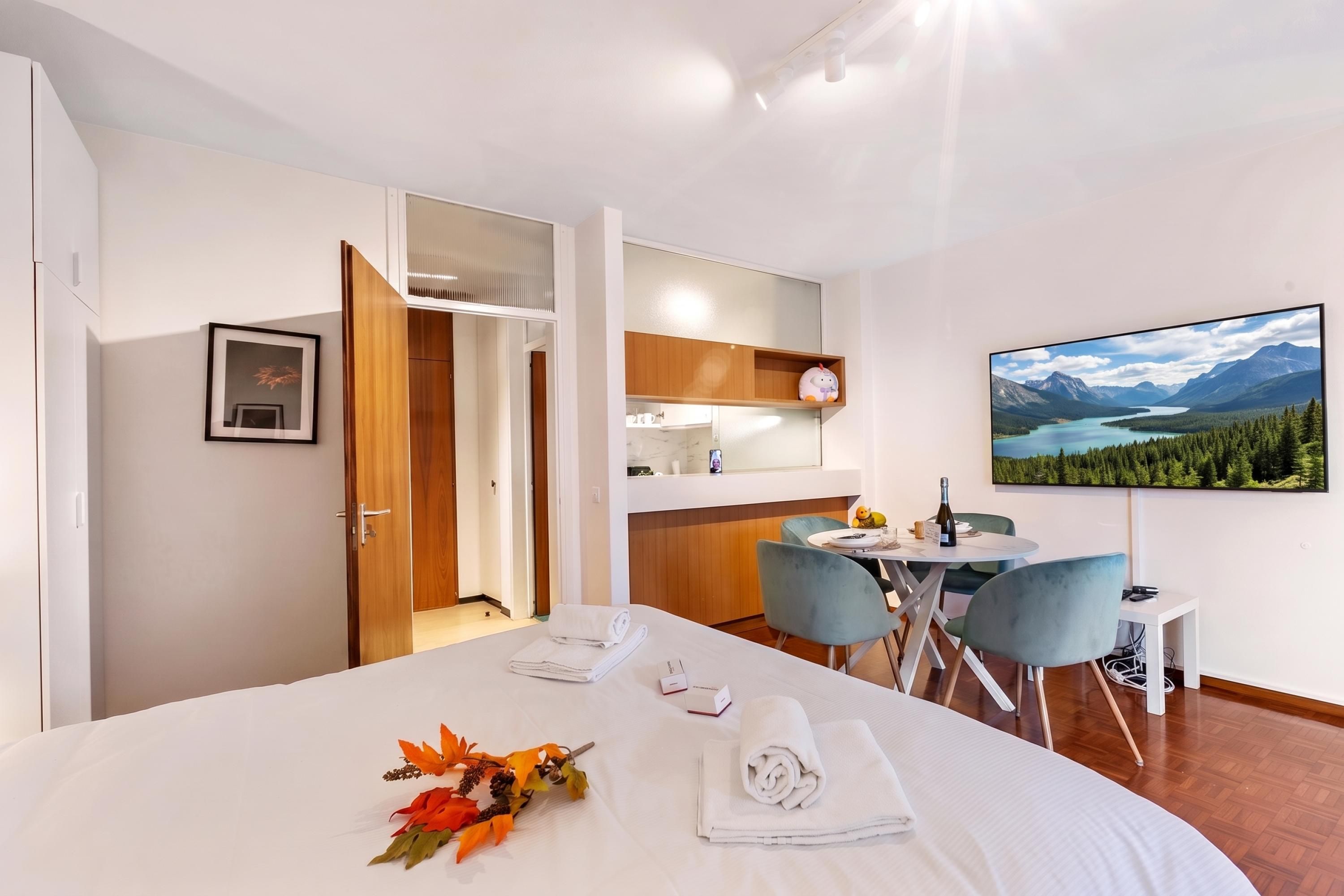 Quarto de hotel com cama, mesa de jantar com cadeiras, área de cozinha e televisão exibindo uma paisagem cênica.