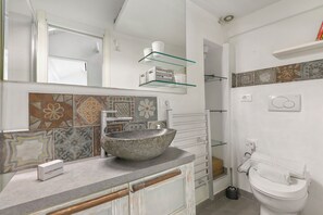 A bathroom with a stone sink, white toilet, and a mirror above the sink. There are patterned tiles on the wall and a shelf with toiletries.
