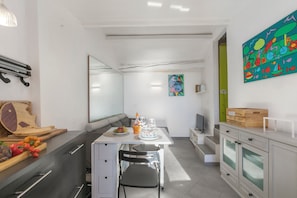 A modern kitchen with a dining table, chairs, and a bench. There is a wooden cutting board with vegetables, a fruit bowl, and a bottle of orange juice. A colorful abstract painting is on the wall, and a wooden box is on the counter. - Un Terrazzo Nella Pigna (Sanremo)