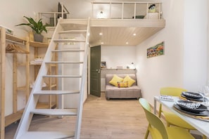 Een loft-achtig vertrek met een houten trap, een bank met kussens, een eettafel met stoelen en een klein balkon.