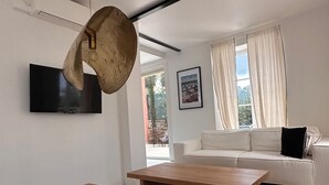 Ein modernes Wohnzimmer mit Flachbildfernseher, zwei Holztischen, einem weißen Sofa und einem großen Fenster mit Vorhängen.