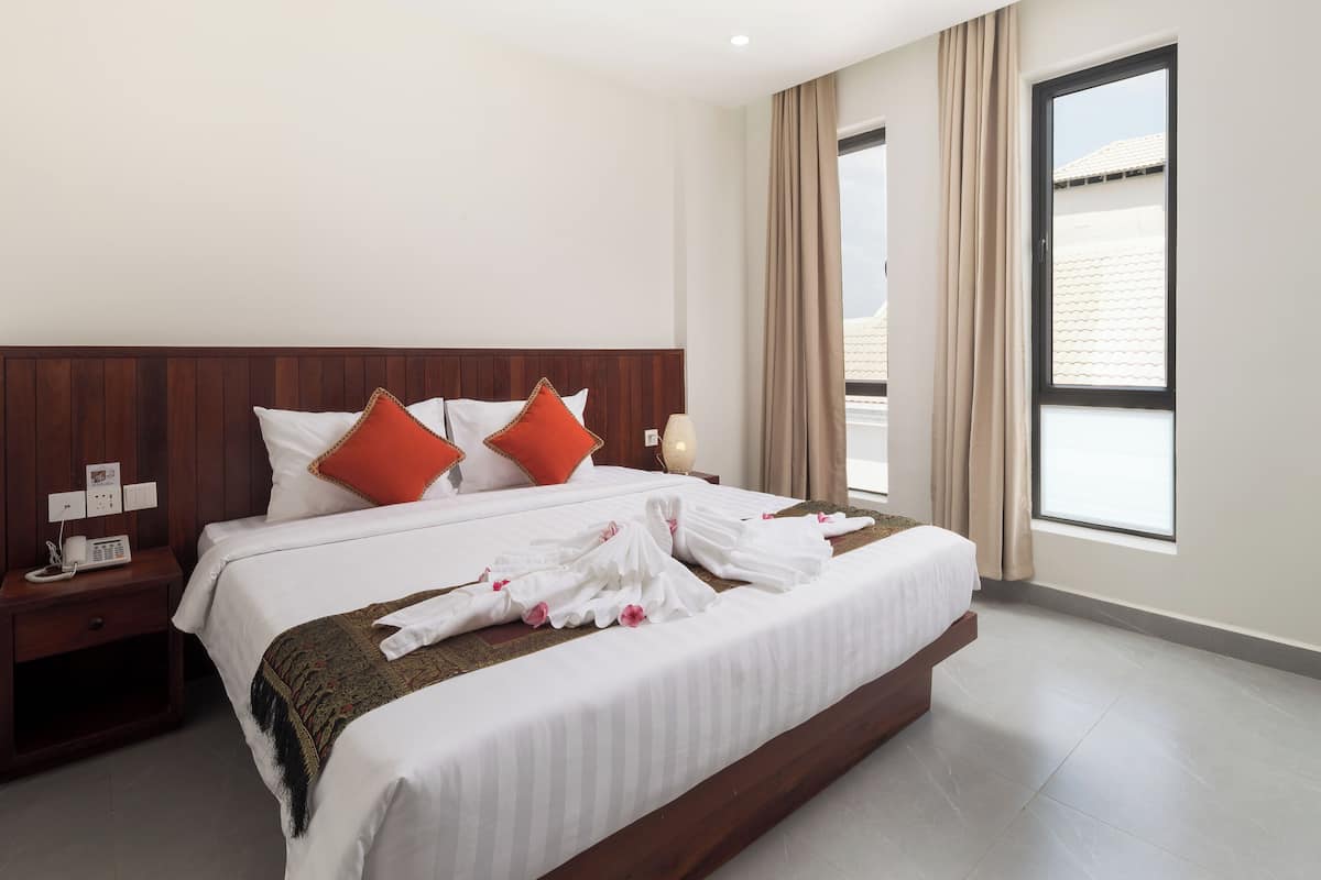 A hotel room with a large bed, wooden headboard, white bedding, and a view of the sky through a window.