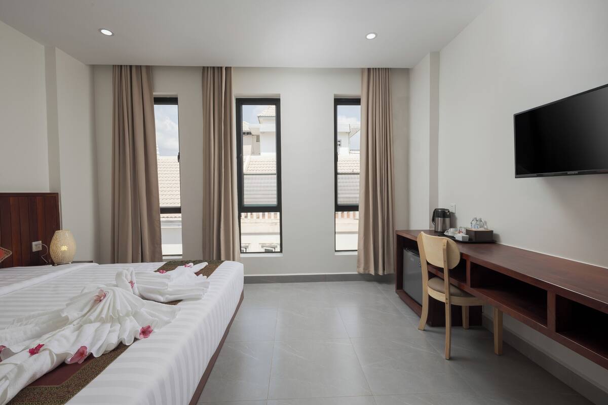 A hotel room with a bed, a desk, a chair, a television, and large windows with curtains.