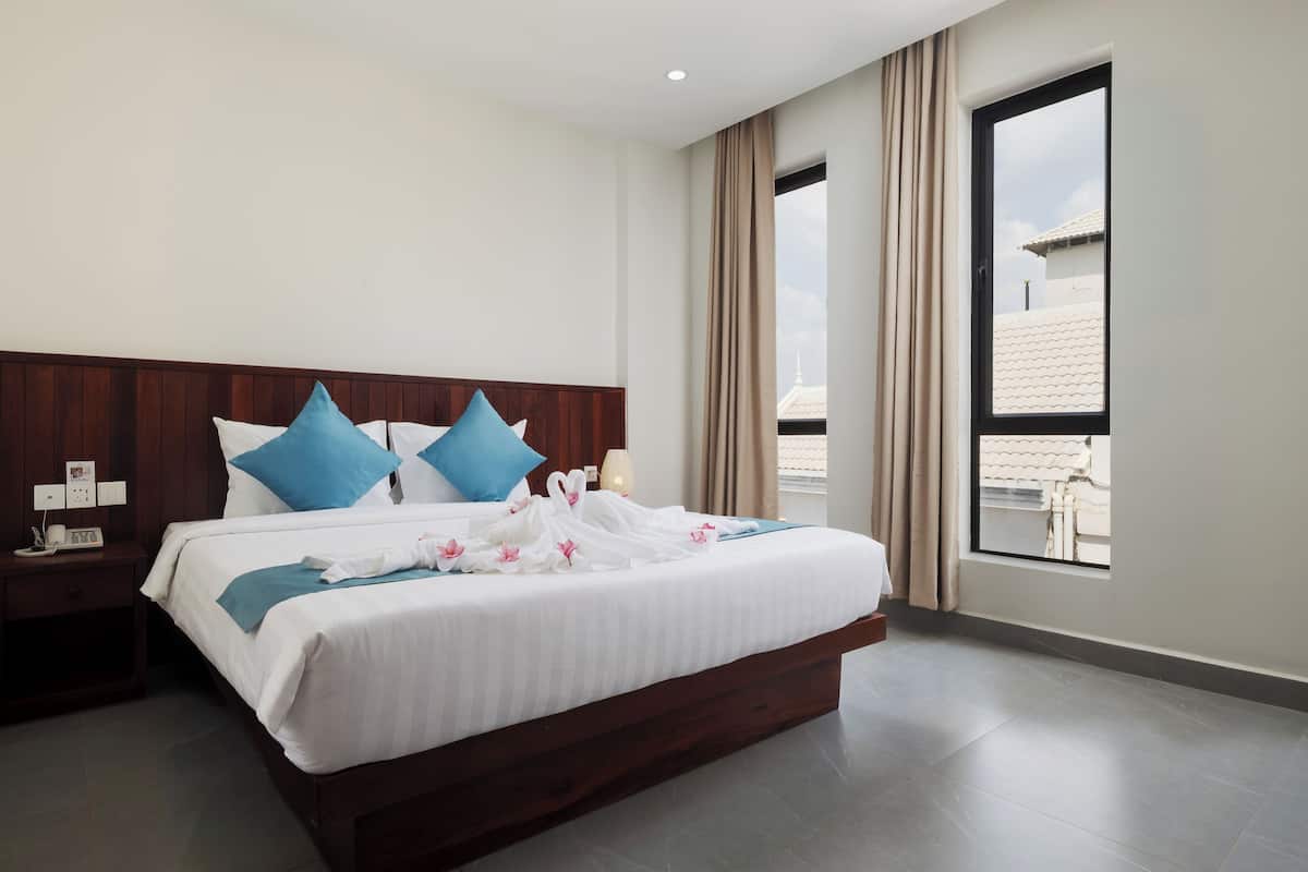 A hotel room with a large bed, a wooden headboard, a nightstand, a window with curtains, and a view of buildings outside.