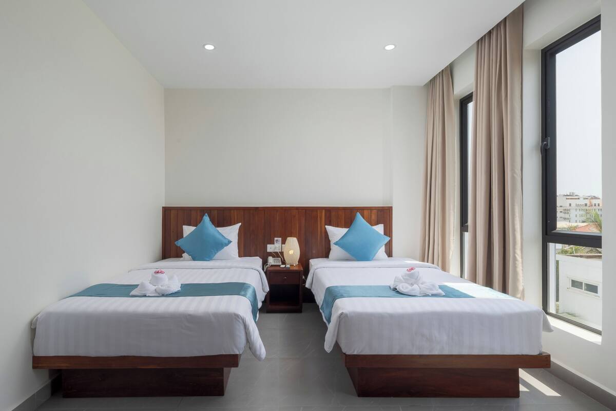 A hotel room with two beds, a wooden headboard, a nightstand with a lamp, and a view of the city through large windows.