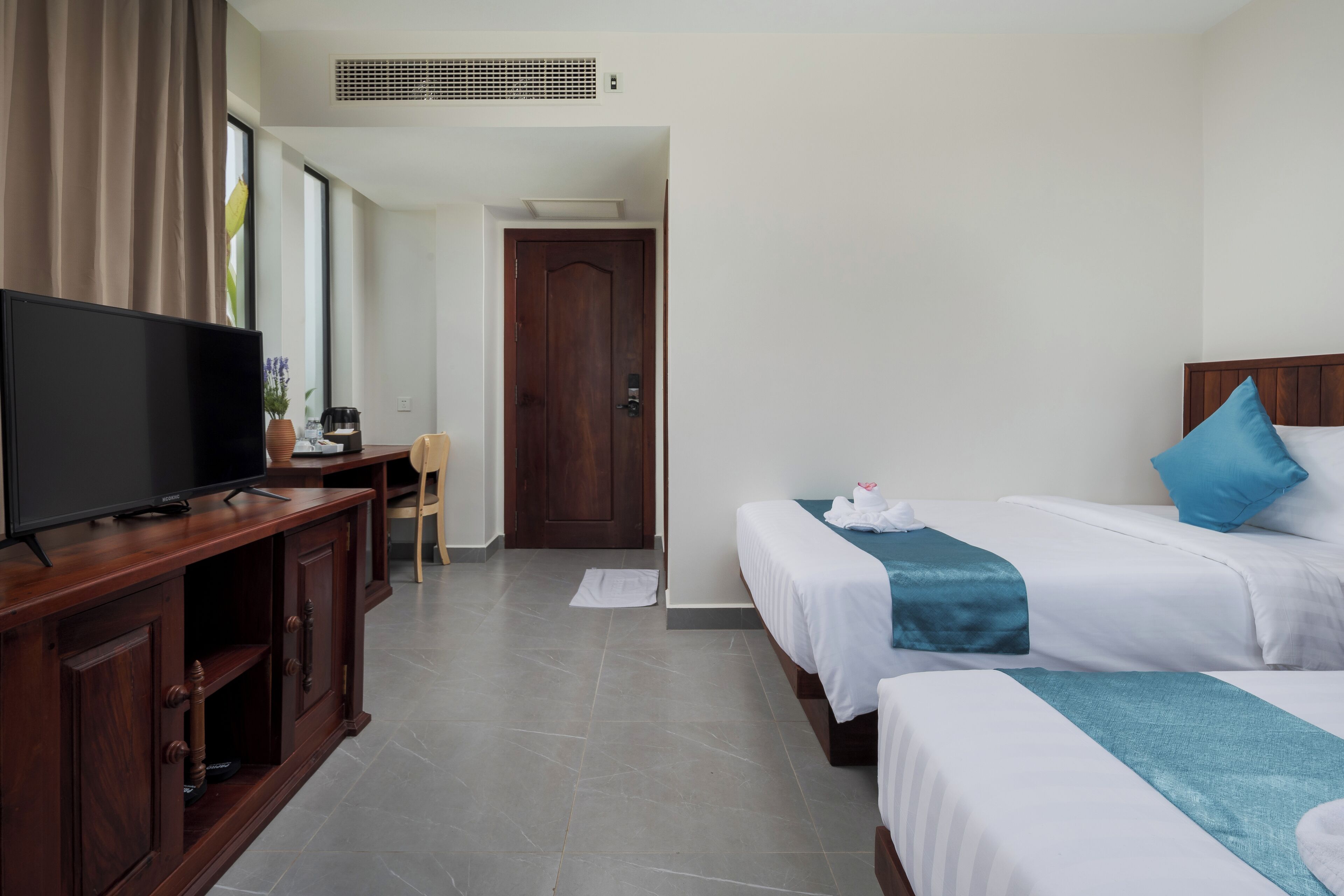 A hotel room with a TV on a wooden cabinet, two beds with white and blue bedding, a wooden door, and a tiled floor.