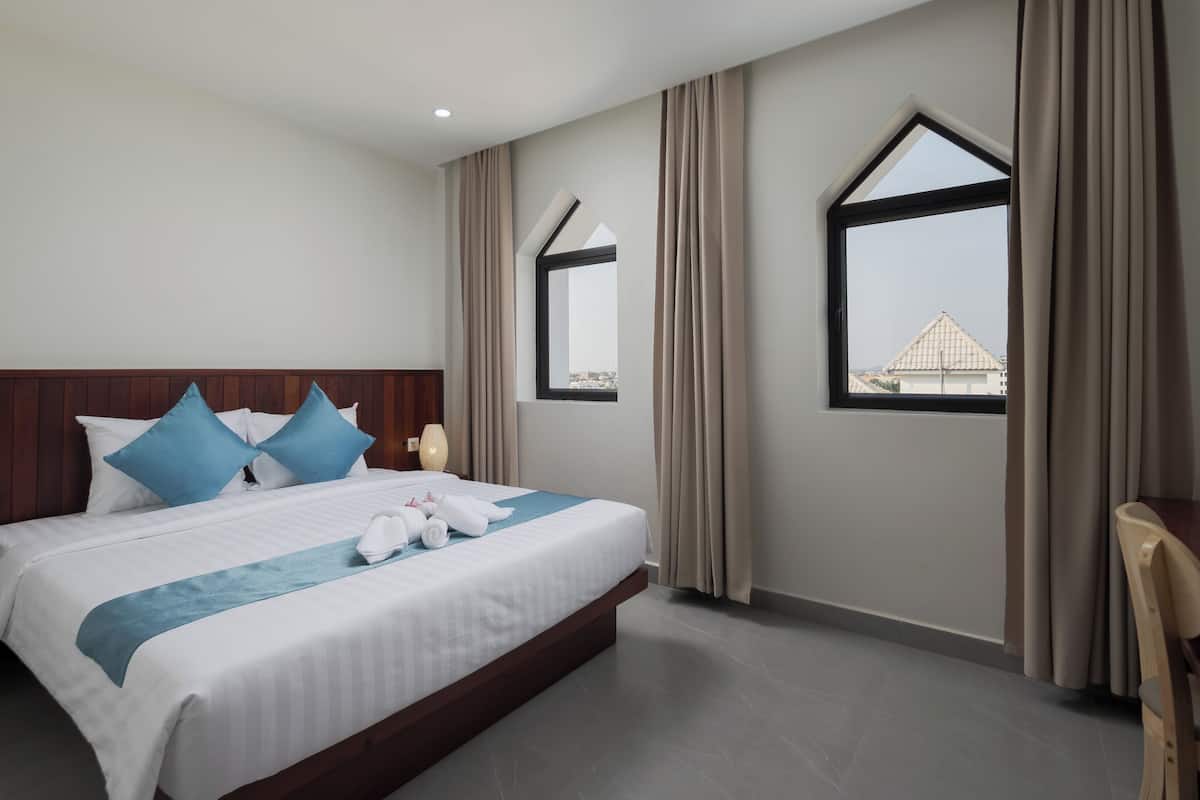 A modern hotel room with a large bed, two windows, and a view of buildings outside.