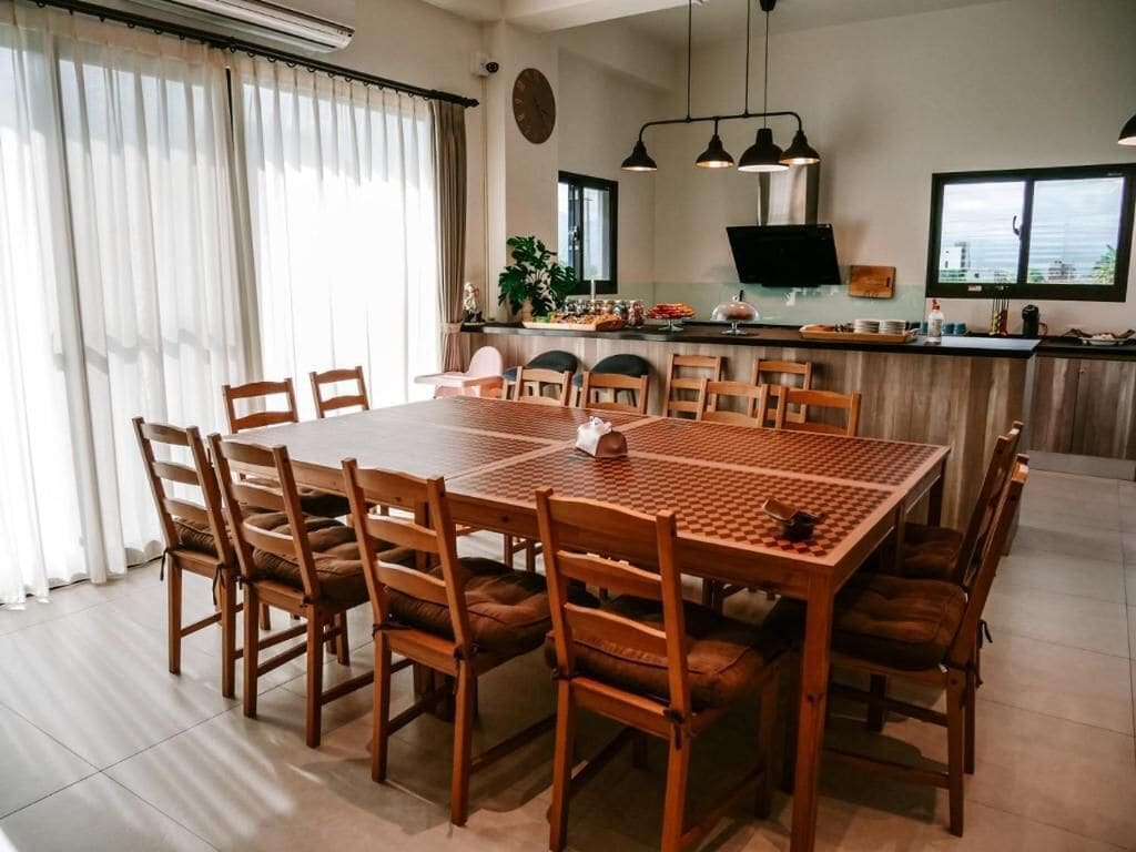 A dining area with a large wooden table and chairs, a kitchen in the background, and a view of the city through large windows.