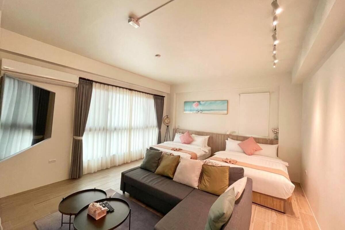 A modern hotel room with a sofa, two beds, a small table, and a large window with curtains.