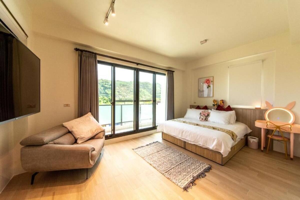 A bedroom with a large bed, a sofa, a mirror, and a view of greenery outside.