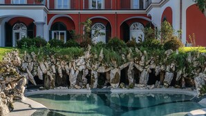 Una piscina con agua cristalina, rodeada por un jardín rocoso y un edificio con paredes rojas y blancas y ventanas de arco.