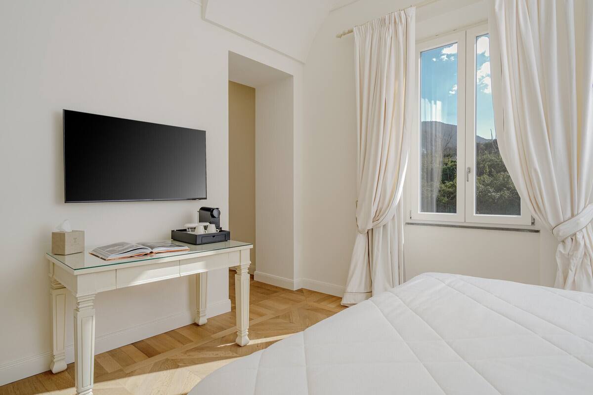 Ett hotellrum med en säng, ett skrivbord med en platt-TV, en kaffebryggare och ett fönster med gardiner.