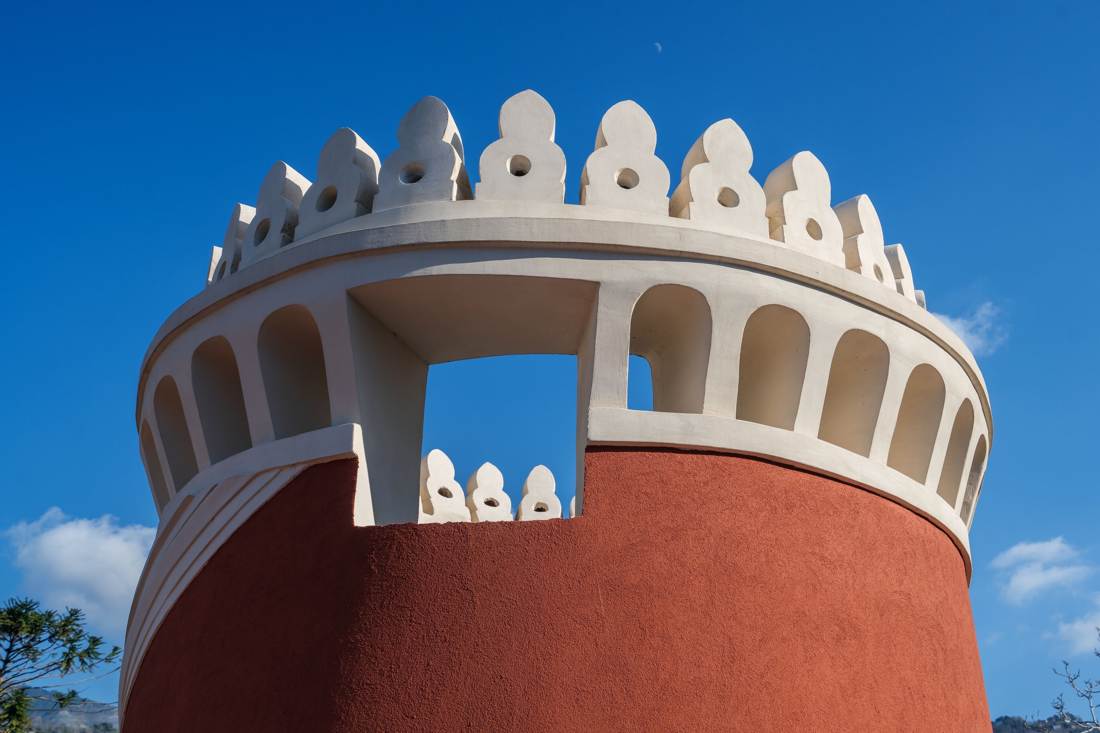 Estrutura circular com topo branco e ornamentado e base vermelha, diante de um céu azul límpido.