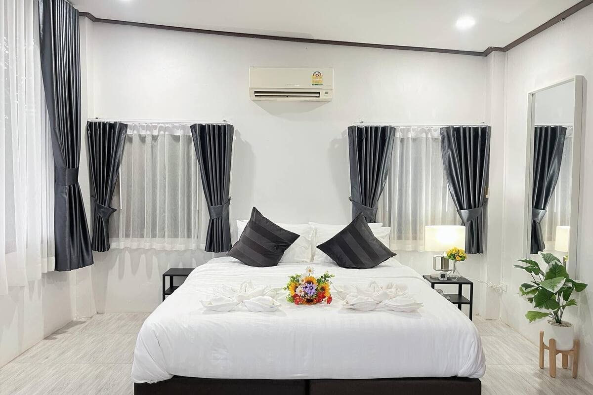 A modern hotel room with a large bed, black curtains, a small table with a lamp, and a potted plant.