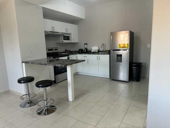 A modern kitchen with white cabinets, a stainless steel refrigerator, a microwave, and a coffee maker. There are two black bar stools at the kitchen island.