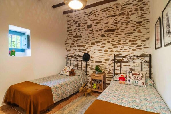 A bedroom with two single beds, a stone wall, a small window, and a nightstand with a lamp.