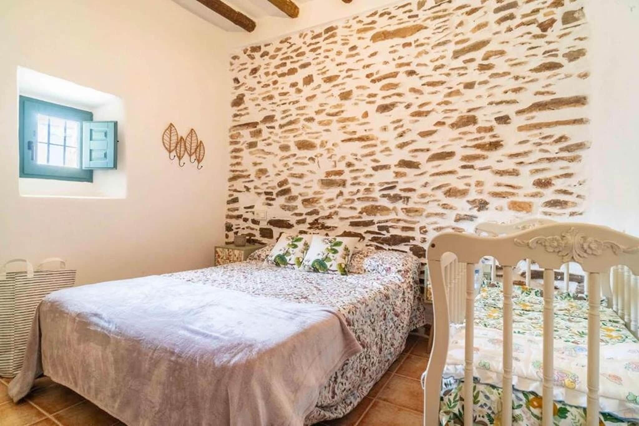 Une chambre avec un mur en pierre, un lit recouvert d’une literie à motifs floraux, un berceau blanc et une fenêtre avec un cadre bleu.