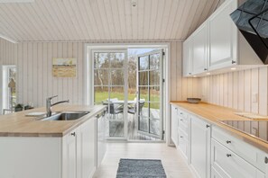 Moderni keittiö, jossa on puinen työtaso, valkoiset kaapit, suuri liukuovi, joka johtaa ulkoterasille, ja riippuvalaisin.