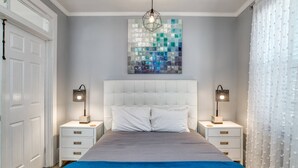 Ein Schlafzimmer mit einem weißen Kopfteil, zwei Nachttischen mit Lampen, einer blauen Bettdecke und einem Mosaikbild an der Wand.